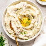 Overhead shot mashed potatoes in a white bowl. Bowl is surrounded by bowls of butter, salt and pepper, and chopped chives.