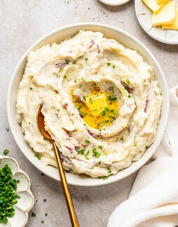Overhead shot mashed potatoes in a white bowl. Bowl is surrounded by bowls of butter, salt and pepper, and chopped chives.