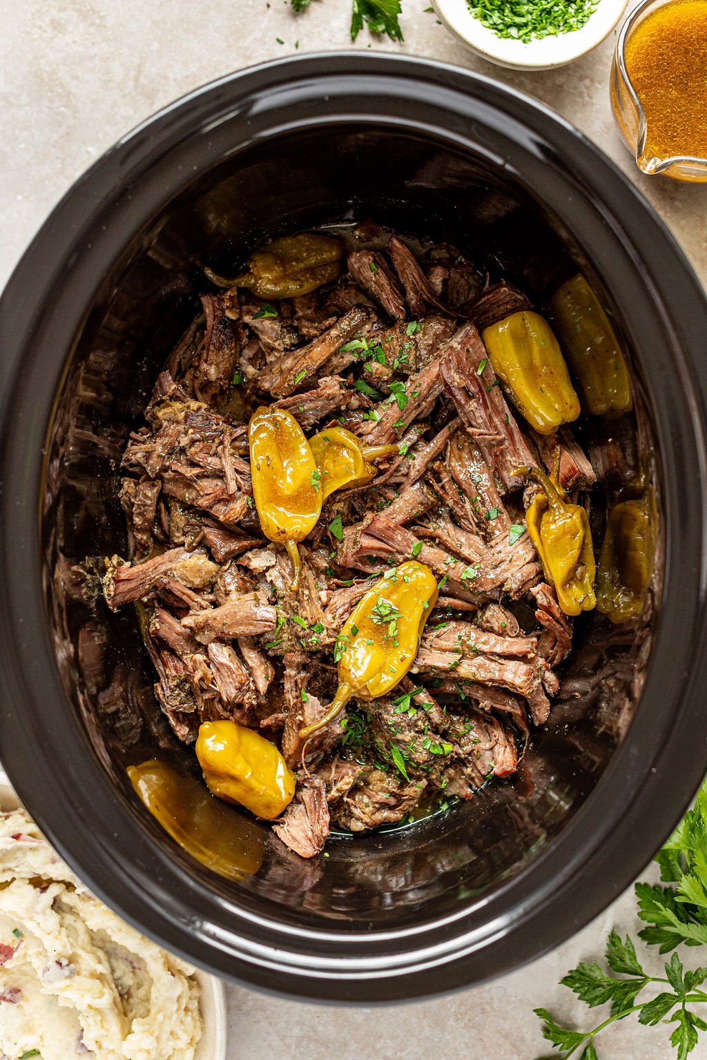Overhead shot of slow cooker with shredded roast beef and pepperoncini peppers. Slow cooker is surrounded by a gravy boat, bowl of chopped parsley, sprigs of fresh parsley, and a bowl of mashed potatoes.