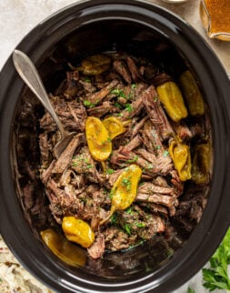 Overhead shot of shredded roast beef being served out of a slow cooker with a serving fork. Slow cooker is surrounded by a gravy boat, bowl of chopped parsley, sprigs of fresh parsley, and a bowl of mashed potatoes.