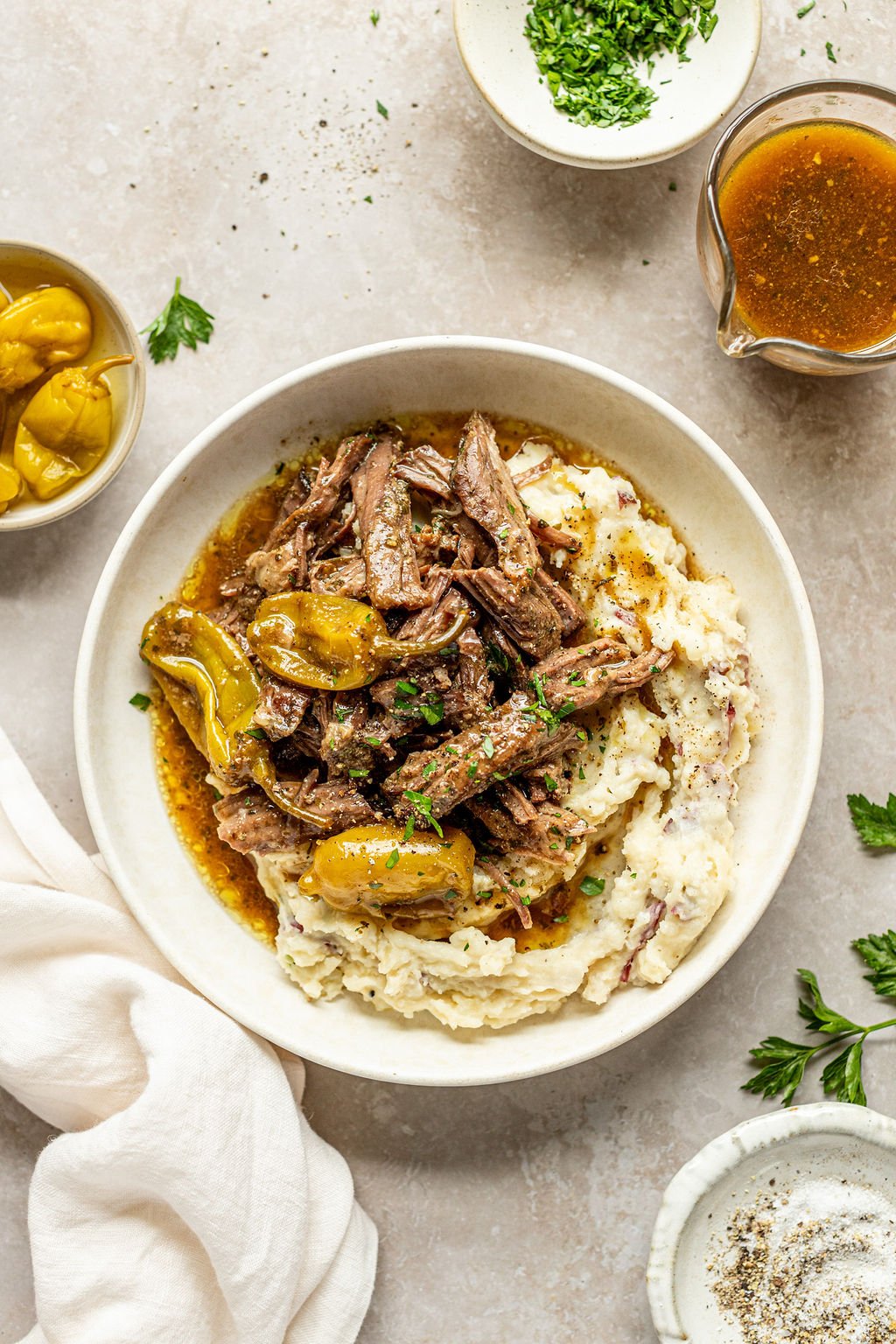 Overhead shot of a white bowl with mashed potatoes and roast beef. Bowl is surrounded by a towel, gravy boat, and bowls or fresh parsley and salt and pepper.