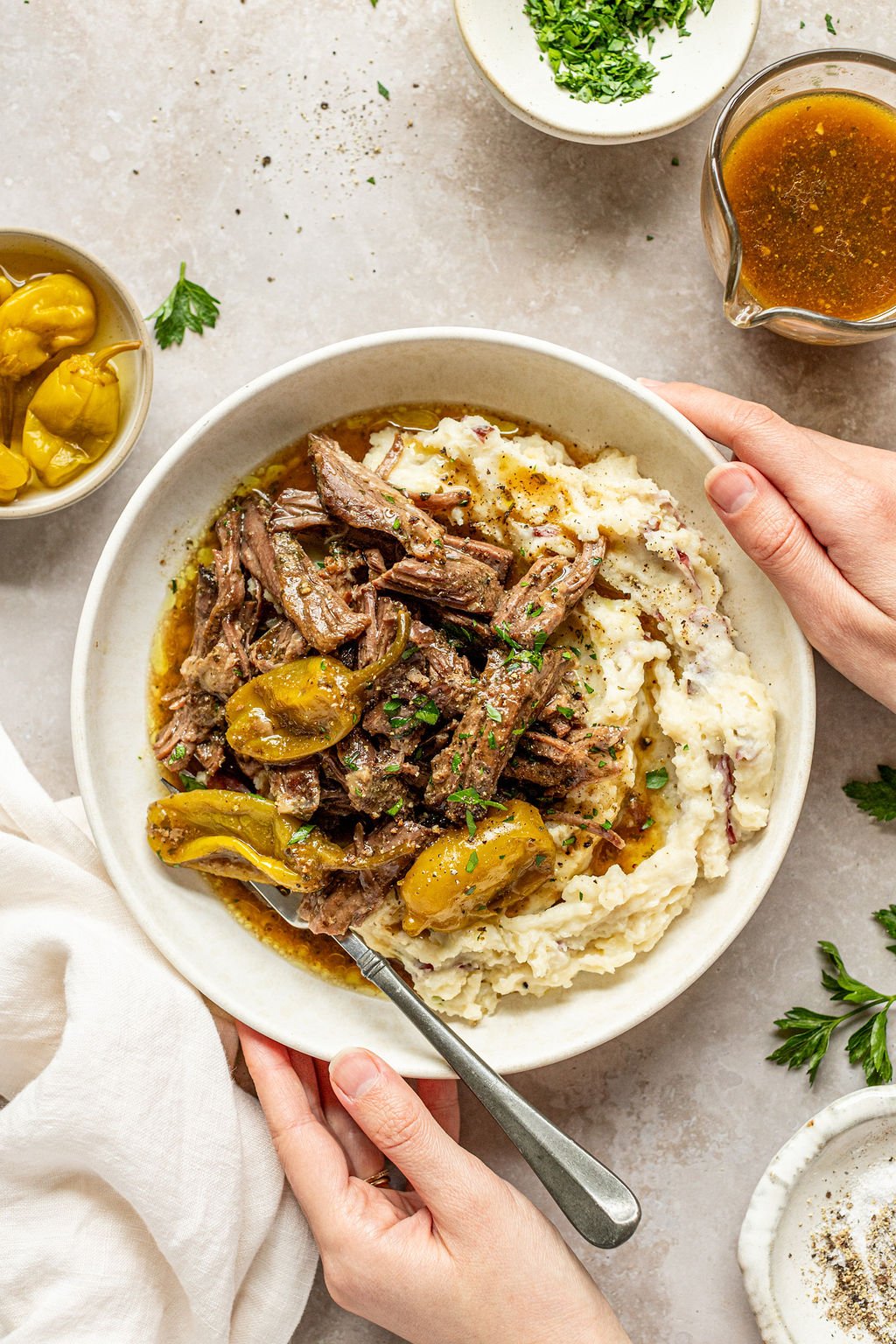 Overhead shot of hands around a white bowl with mashed potatoes and roast beef. Bowl is surrounded by a towel, gravy boat, and bowls or fresh parsley and salt and pepper.