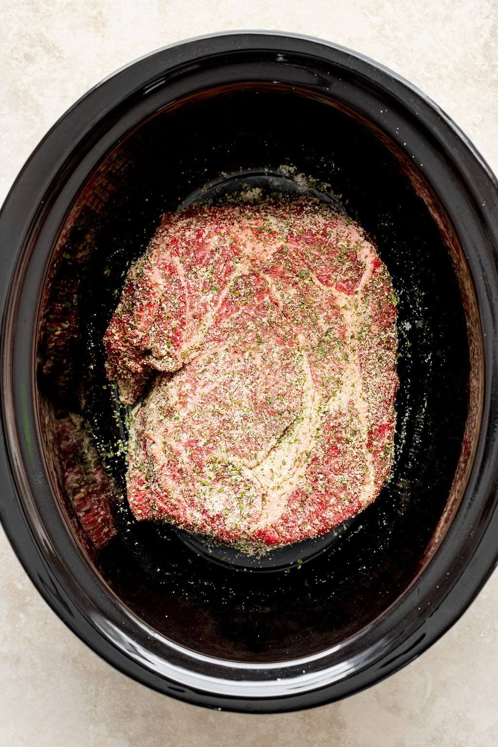 Overhead shot of raw raw chuck roast with seasonings in a slow cooker.