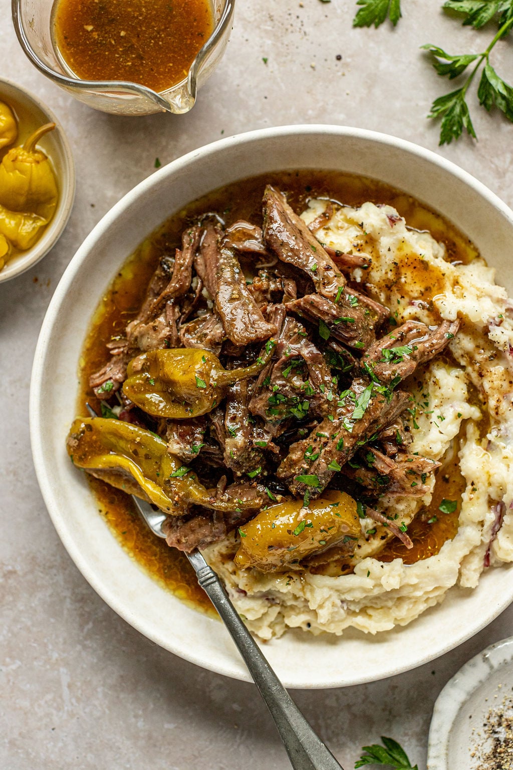 Close-up overhead shot of a white bowl with mashed potatoes and roast beef. Bowl is surrounded by a towel, gravy boat, sprigs of fresh parsley, and a bowl of salt and pepper.