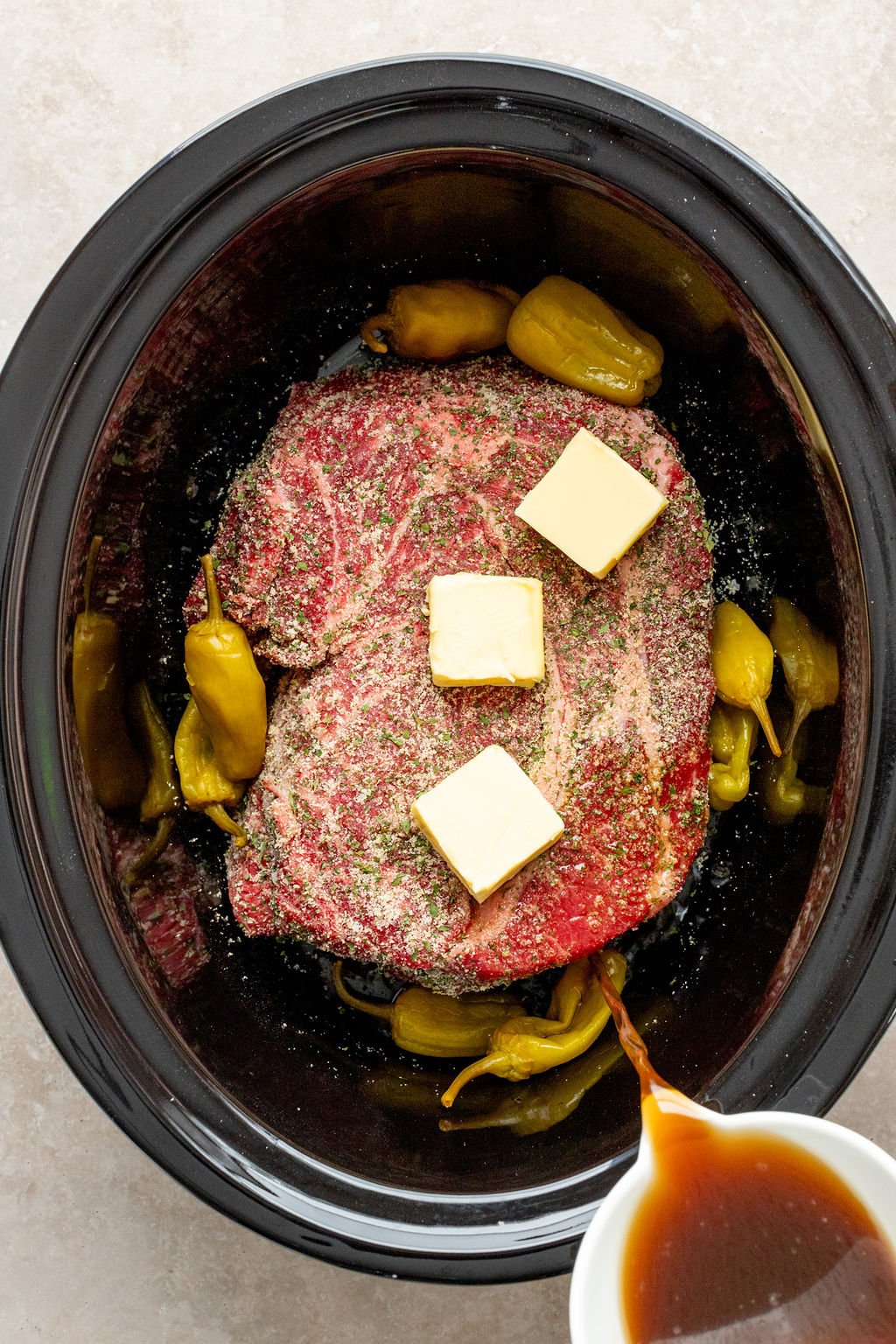 Overhead shot of a slow cooker with raw chuck roast covered in seasonings and butter, surrounded by pepperoncini. Broth is being poured into the slow cooker.