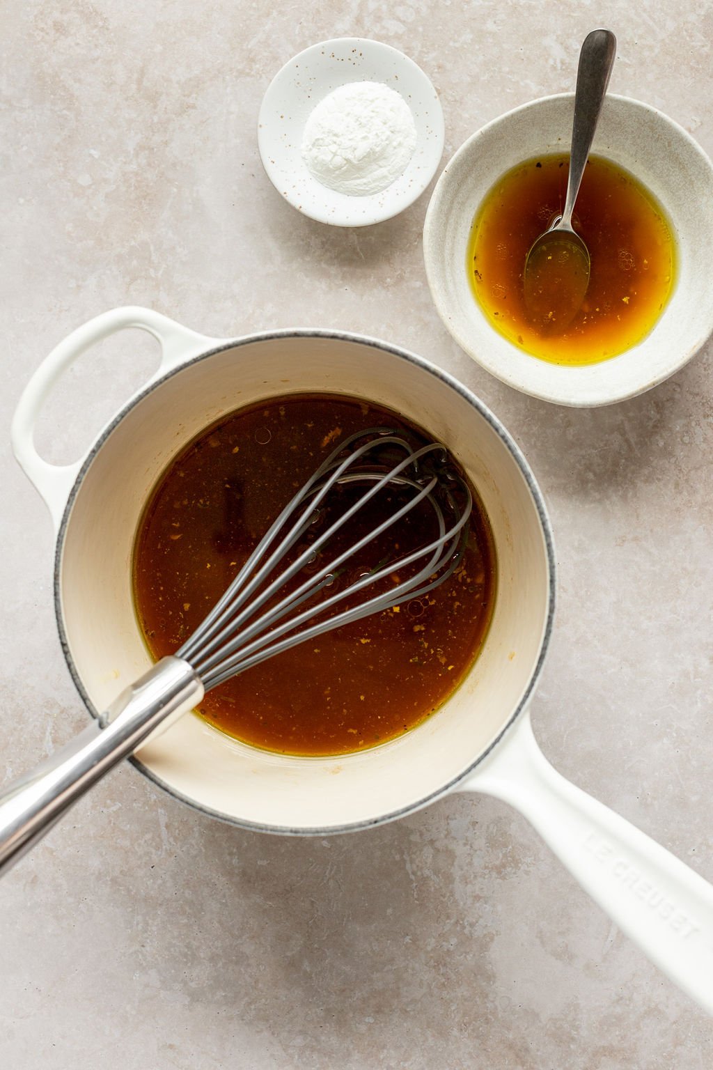 Overhead shot of a small white sauce pan with broth and a whisk in it. Saucepan is surrounded by a small white bowl with broth and a spoon and a pinch bowl with cornstarch.