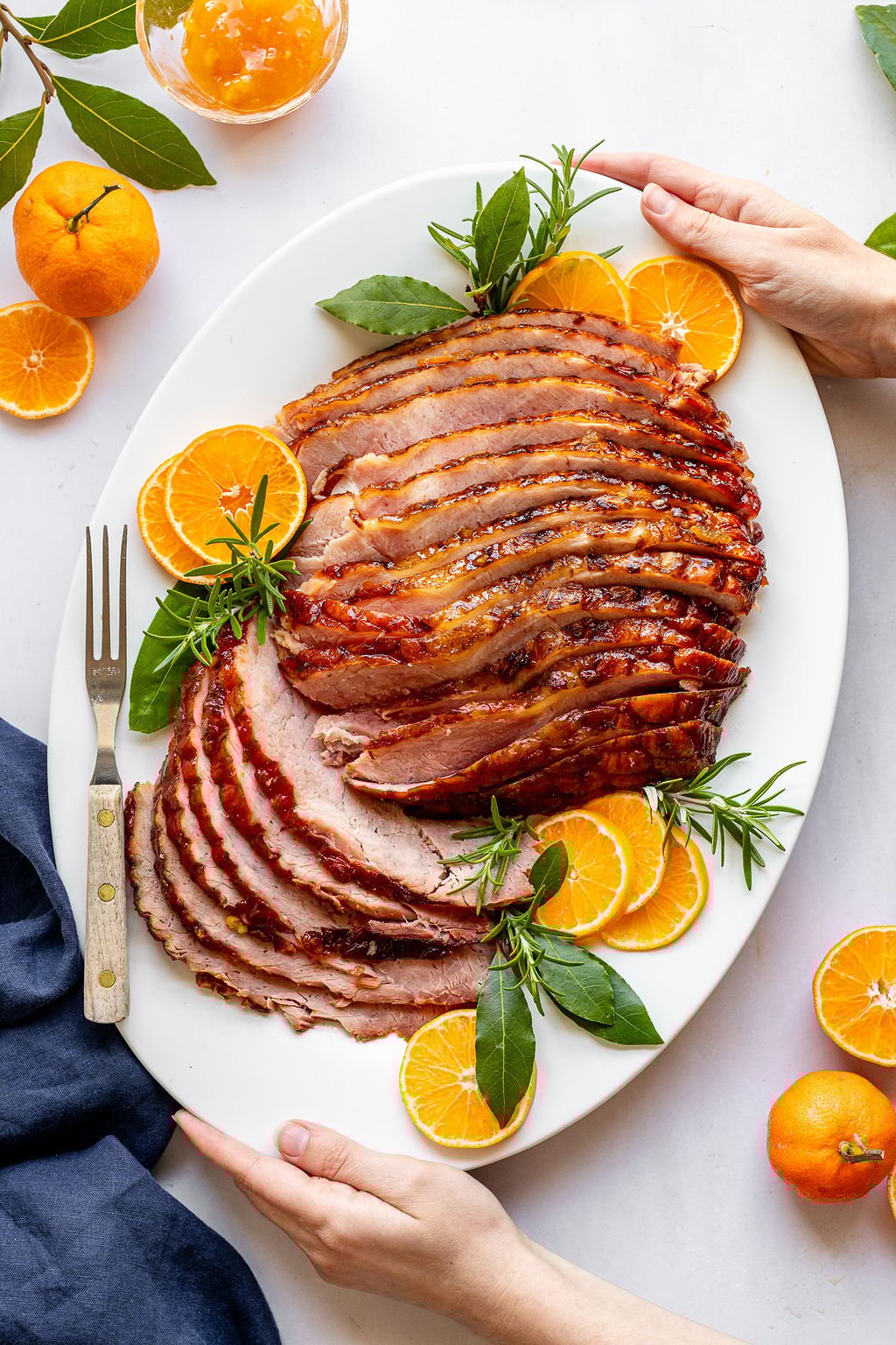 Overhead shot of a white oval serving plate with a serving fork and sliced ham surrounded by orange slices and fresh herbs. Hands are on either side of the plate.