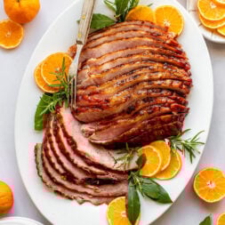 Overhead shot of a white oval serving plate with a serving fork and sliced ham surrounded by orange slices and fresh herbs. Plate is surrounded by oranges, herbs, white serving plates, and a bowl of orange marmalade.