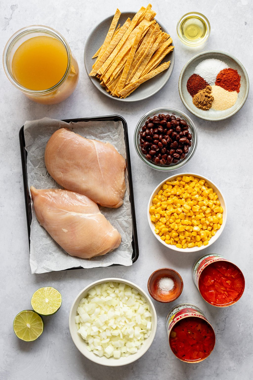 Overhead shot of the following ingredients: broth, tortilla strips, oil, spices, black beans, corn, canned tomatoes, salt, diced onion, limes, and raw chicken breasts.