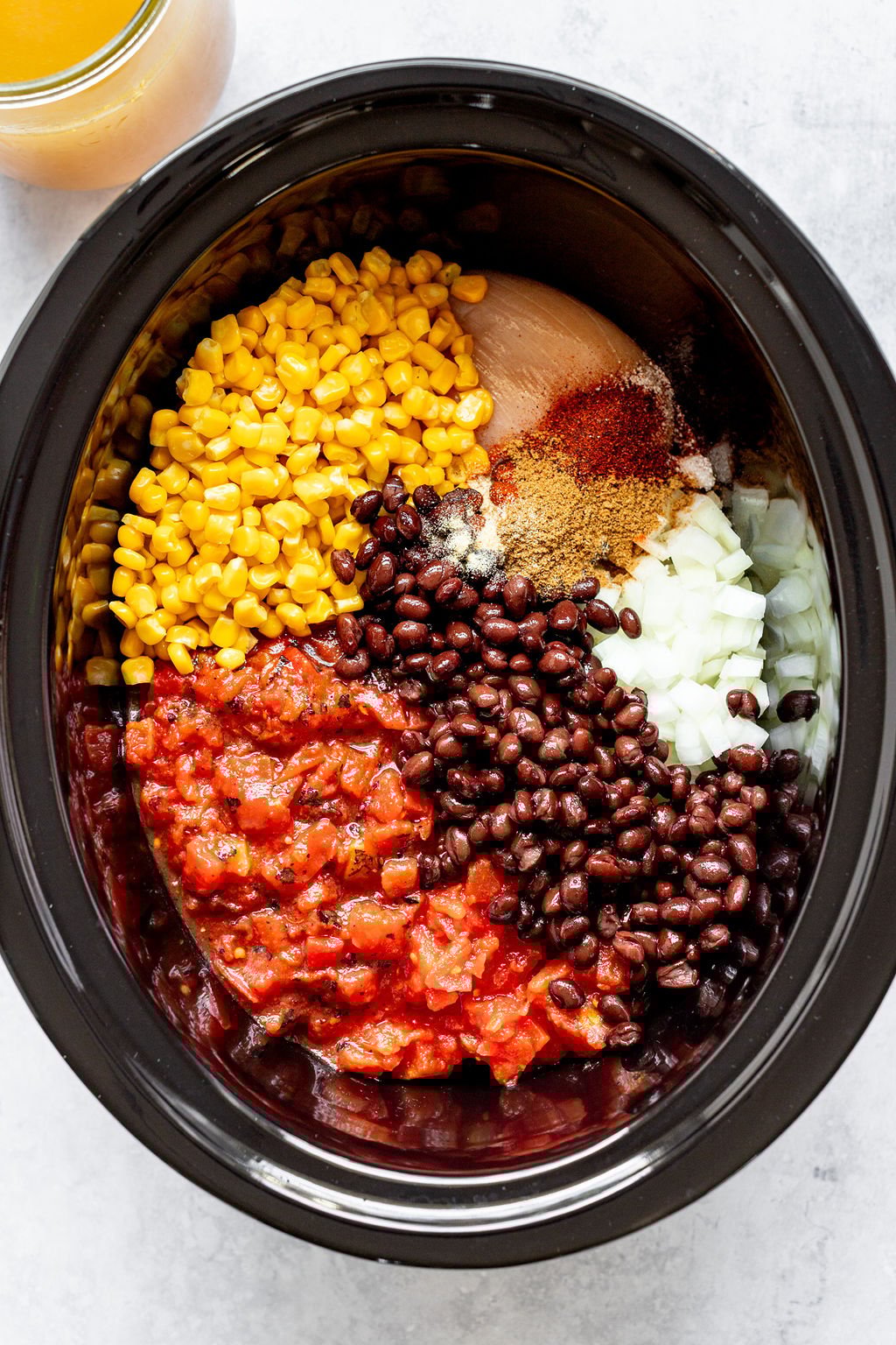 Overhead shot of slow cooker with roasted tomatoes, black beans, corn, spices, raw chicken breast, and chopped onion. A jar of broth is off to the side.