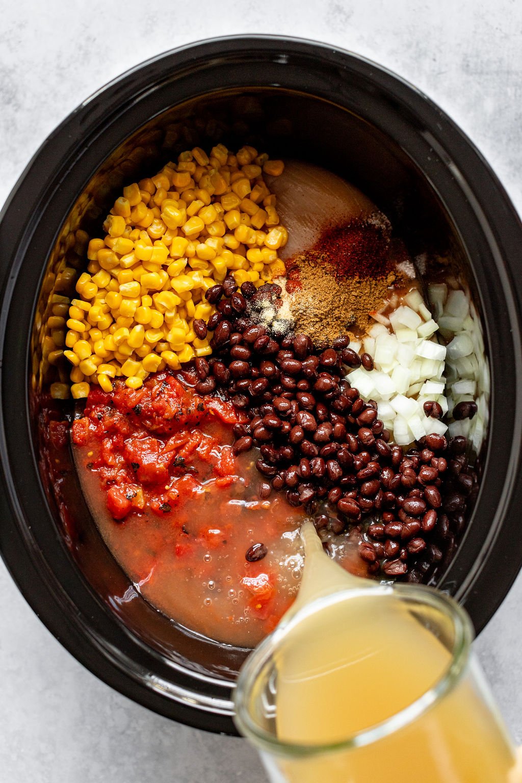 Overhead image of pouring broth into a crock pot of roasted tomatoes, corn, diced onion, black beans, spices, and raw chicken breast.