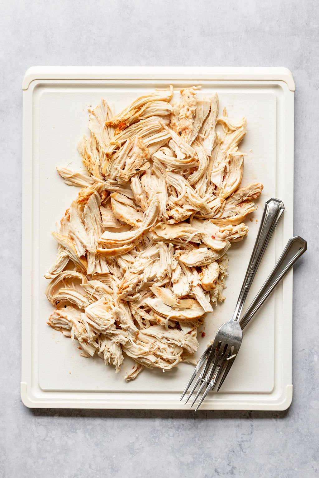 Overhead shot of shredded cooked chicken on a white cutting board with forks on the side.