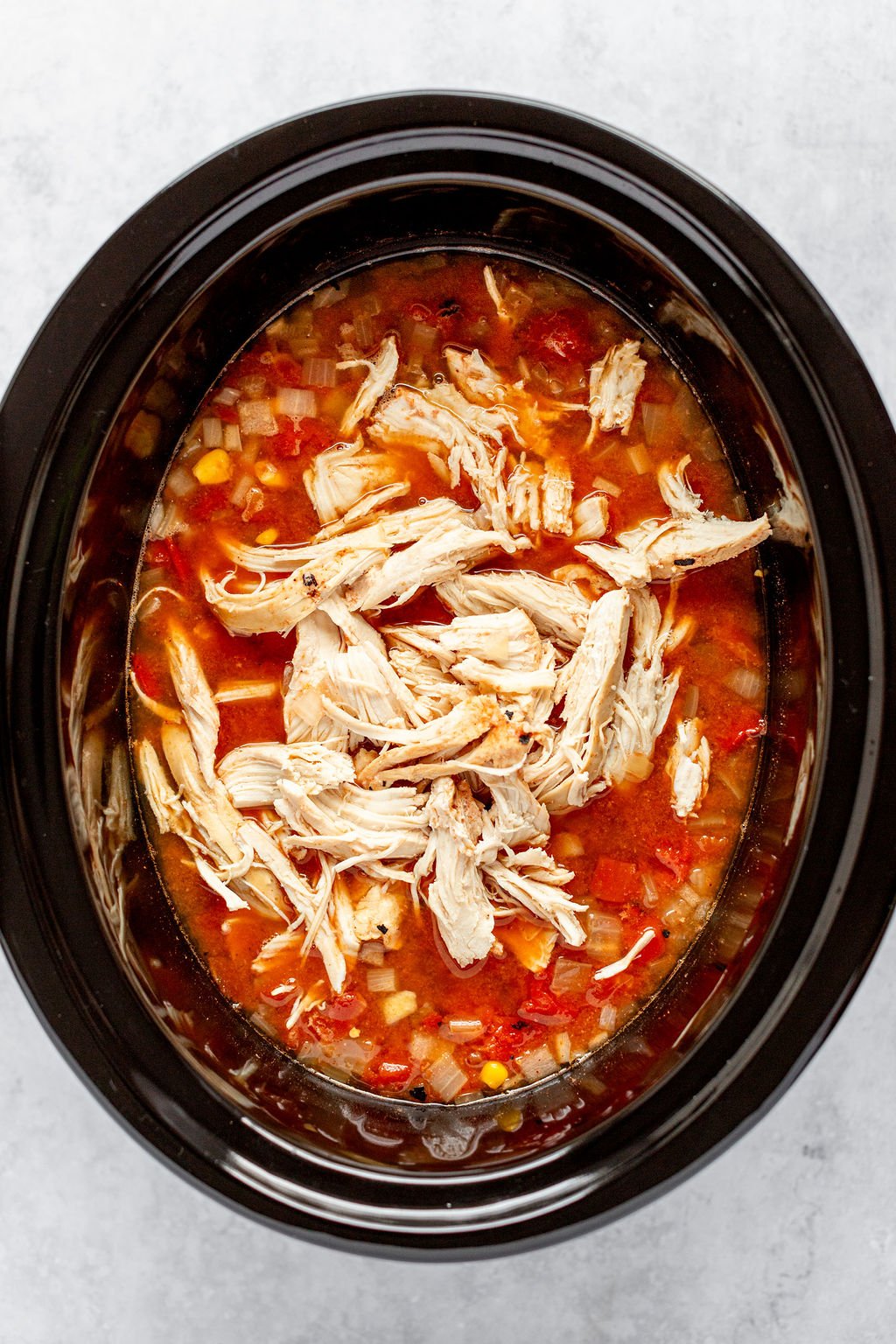 Overhead image of a slow cooker with red broth, corn, and diced onions, and shredded cooked chicken on top.
