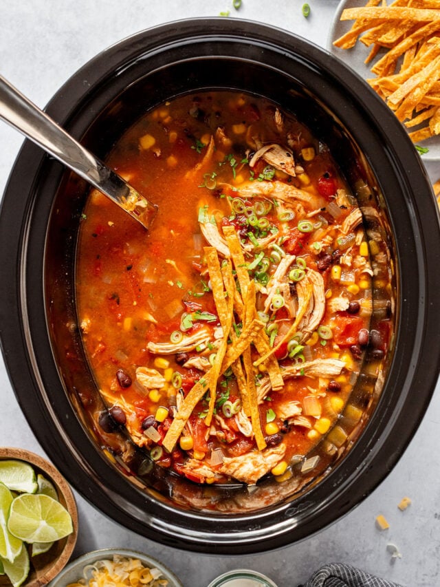 Overhead shot of a ladle in a crockpot filled with broth with beans, corn, shredded chicken, and tortilla strips. Crockpot is surrounded by bowls of lime wedges, shredded cheese, tortilla strips, sour cream, and green onion.