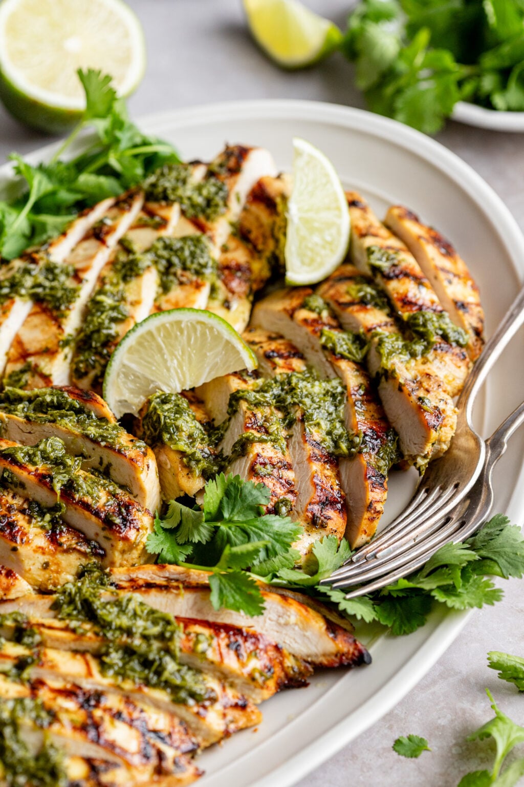 Side view of of white oval platter with two forks, sliced chicken breasts with green sauce, lime wedges, and fresh cilantro.