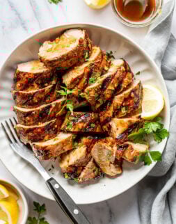 Overhead shot of a white bowl with a serving fork and sliced, grilled pork tenderloin topped with fresh parsley. Bowl is surrounded by lemon wedges, blue towel, and a jar of red sauce.