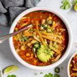 Overhead shot of white bowl with red soup topped with avocado, jalapenos, cilantro, green onions, and tortilla strips. Bowl is surrounded by a grey towel, limes, and a bowl of sliced avocado and jalapenos.