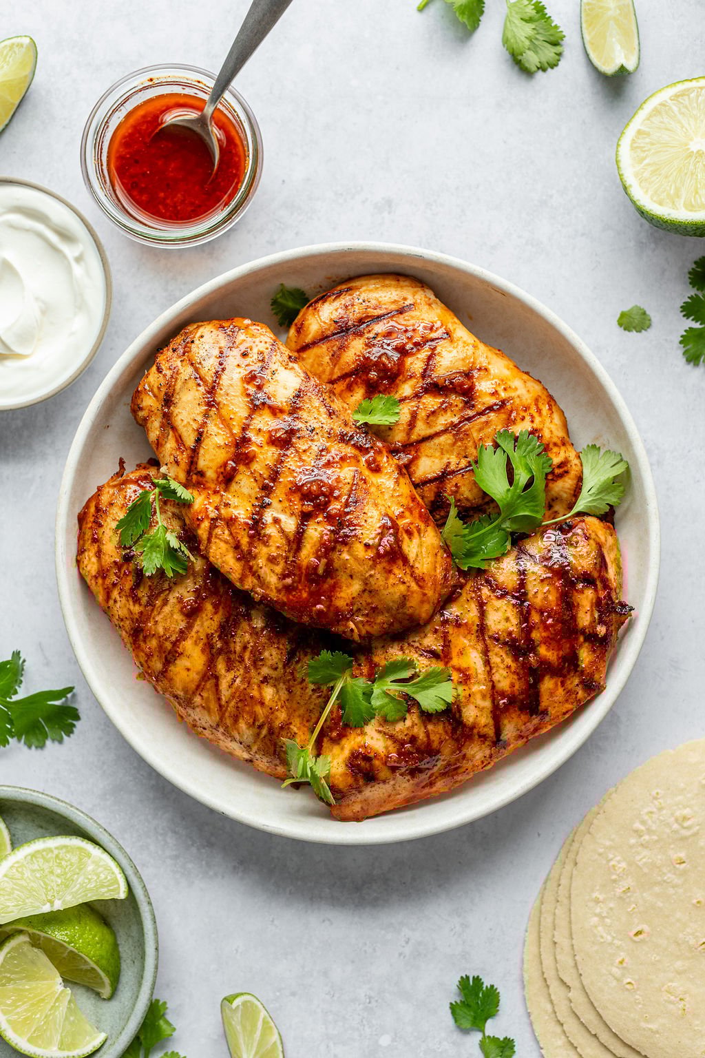 Overhead shot of a white plate with grilled chicken breasts covered in red sauce and fresh cilantro. Plate is surrounded by bowls of sour cream, lime wedges, and red sauce, lime half, cilantro sprigs, and corn tortillas.