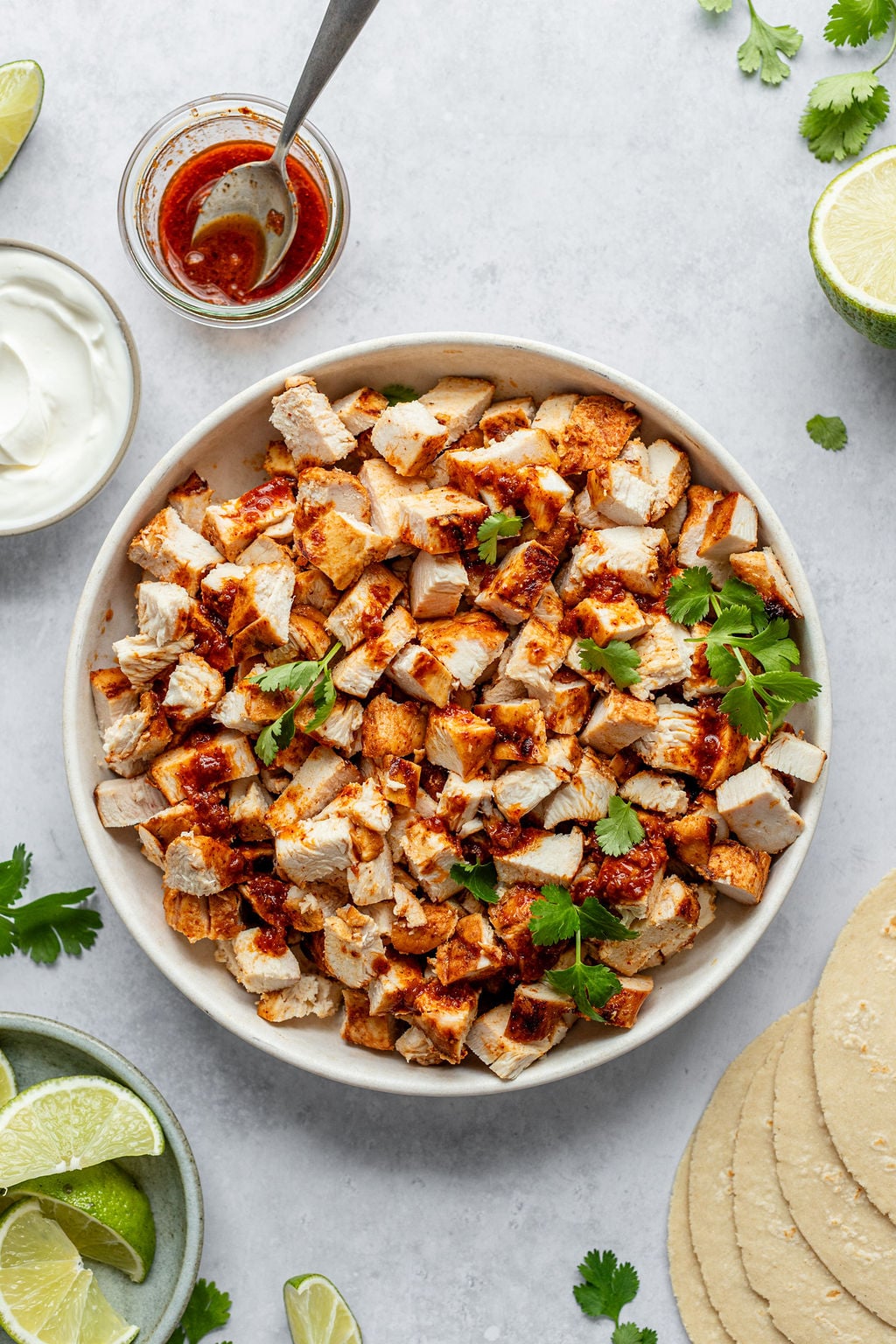 Overhead shot of a large white bowl with diced grilled chicken topped with cilantro sprigs. Bowl is surrounded by sliced limes, a jar of red sauce, and corn tortillas.