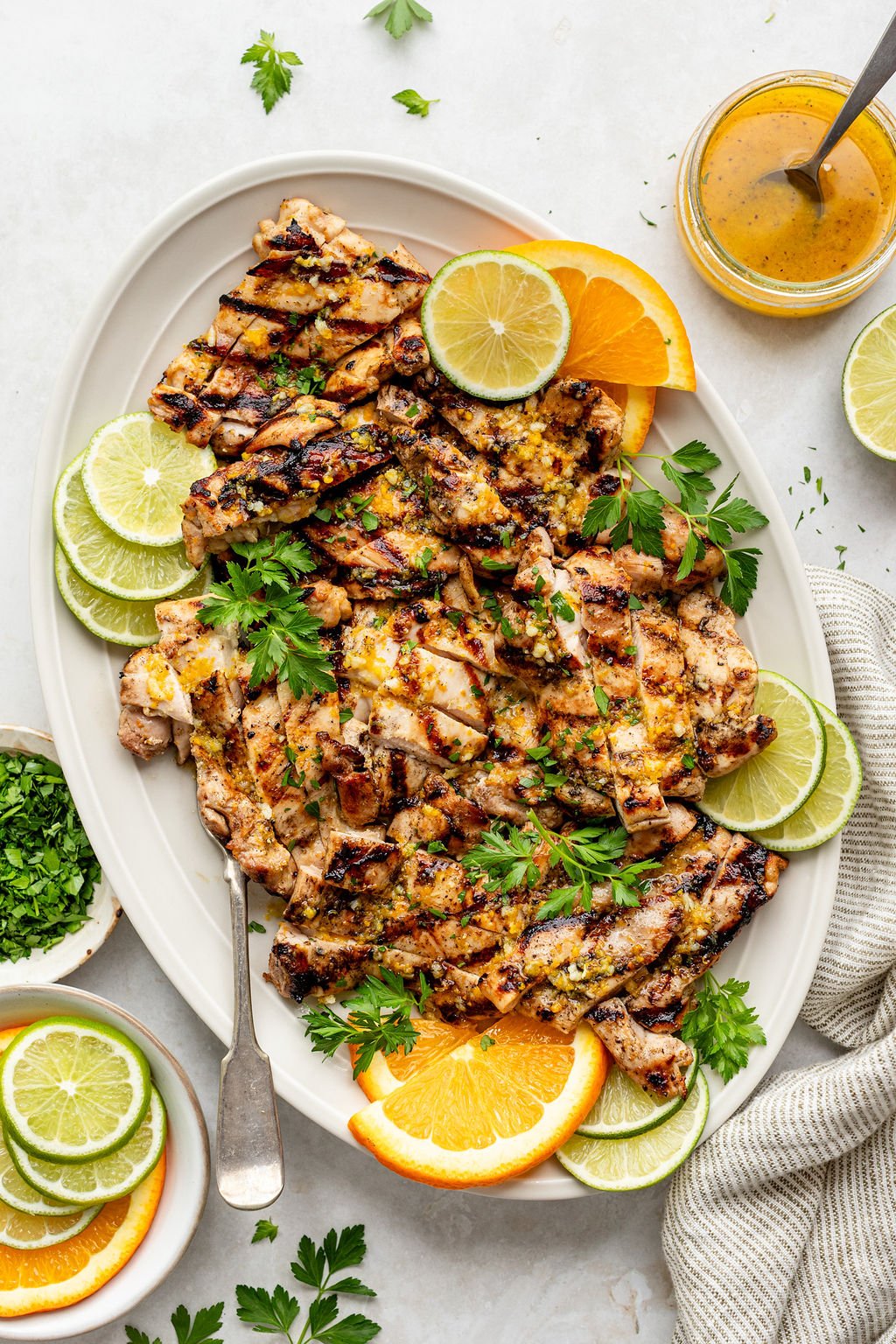 Overhead shot of a white platter with sliced, grilled chicken; a serving fork; fresh herbs; and orange and lime slices. Platter is surrounded by small bowls of lime and orange slices and chopped herbs, a striped towel, and a jar of yellow sauce.