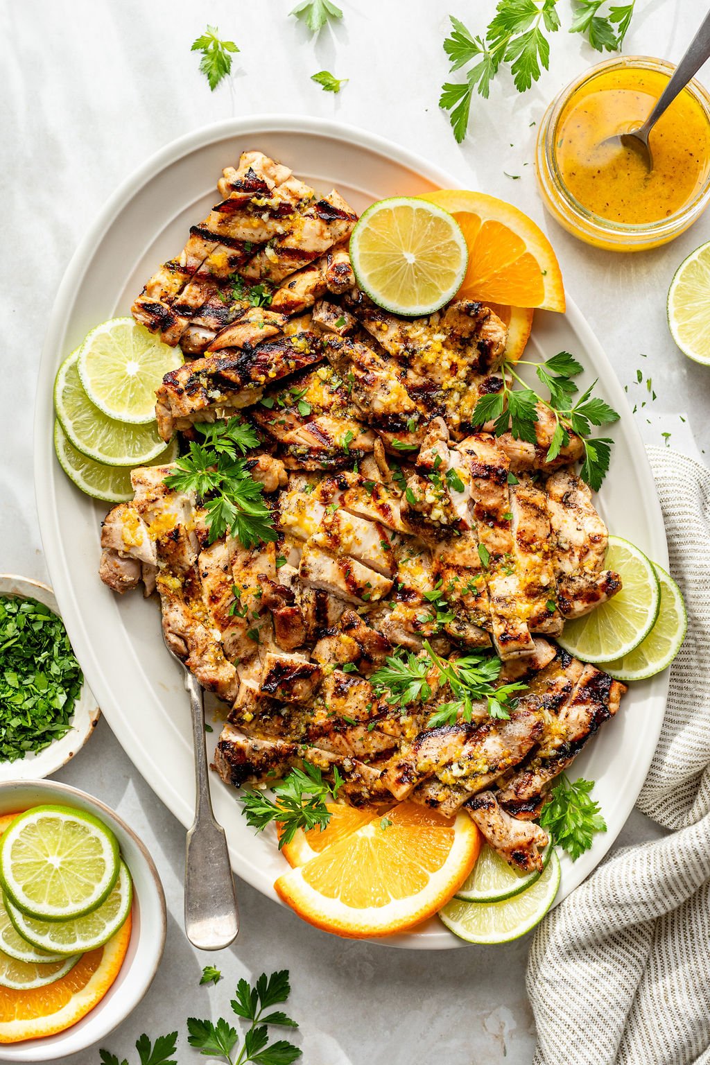 Overhead shot of a white platter with sliced, grilled chicken; a serving fork; fresh herbs; and orange and lime slices. Platter is surrounded by small bowls of lime and orange slices and chopped herbs, a striped towel, and a jar of yellow sauce.