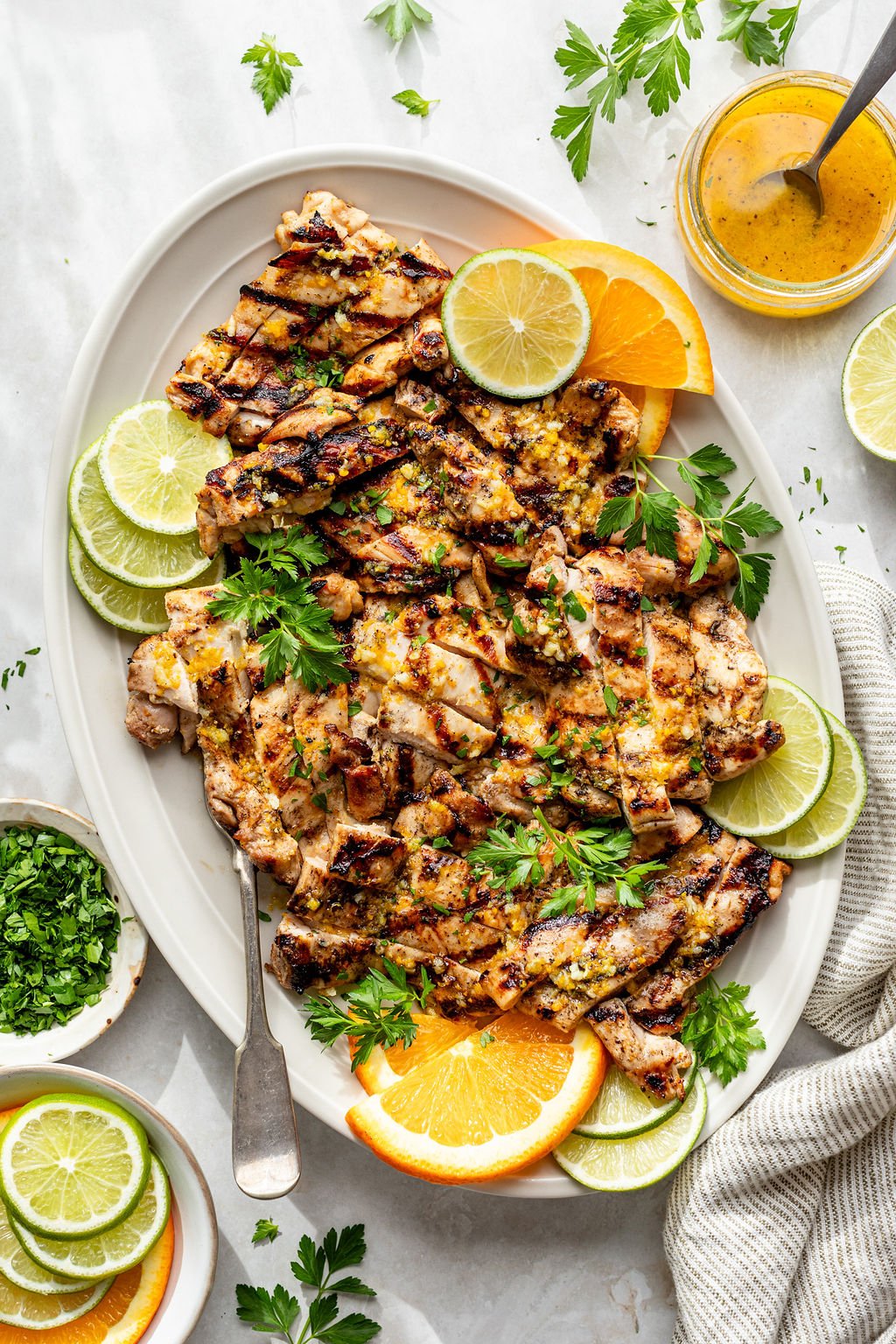 Overhead shot of a white platter with sliced, grilled chicken; a serving fork; fresh herbs; and orange and lime slices. Platter is surrounded by small bowls of lime and orange slices and chopped herbs, a striped towel, and a jar of yellow sauce.