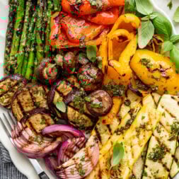 Close-up overhead shot of a white serving platter with a serving fork and grilled mushrooms, zucchini, yellow squash, bell peppers, red onion, and asparagus topped with oil and herb mixture.