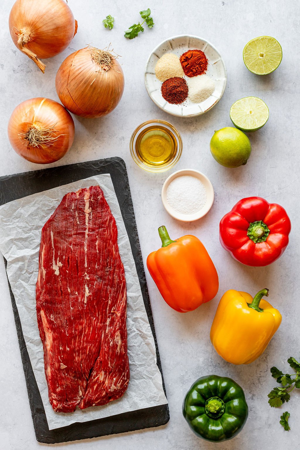 Overhead shot of the following ingredients: steak, onions, spices, oil, limes, salt, bell peppers, and cilantro.