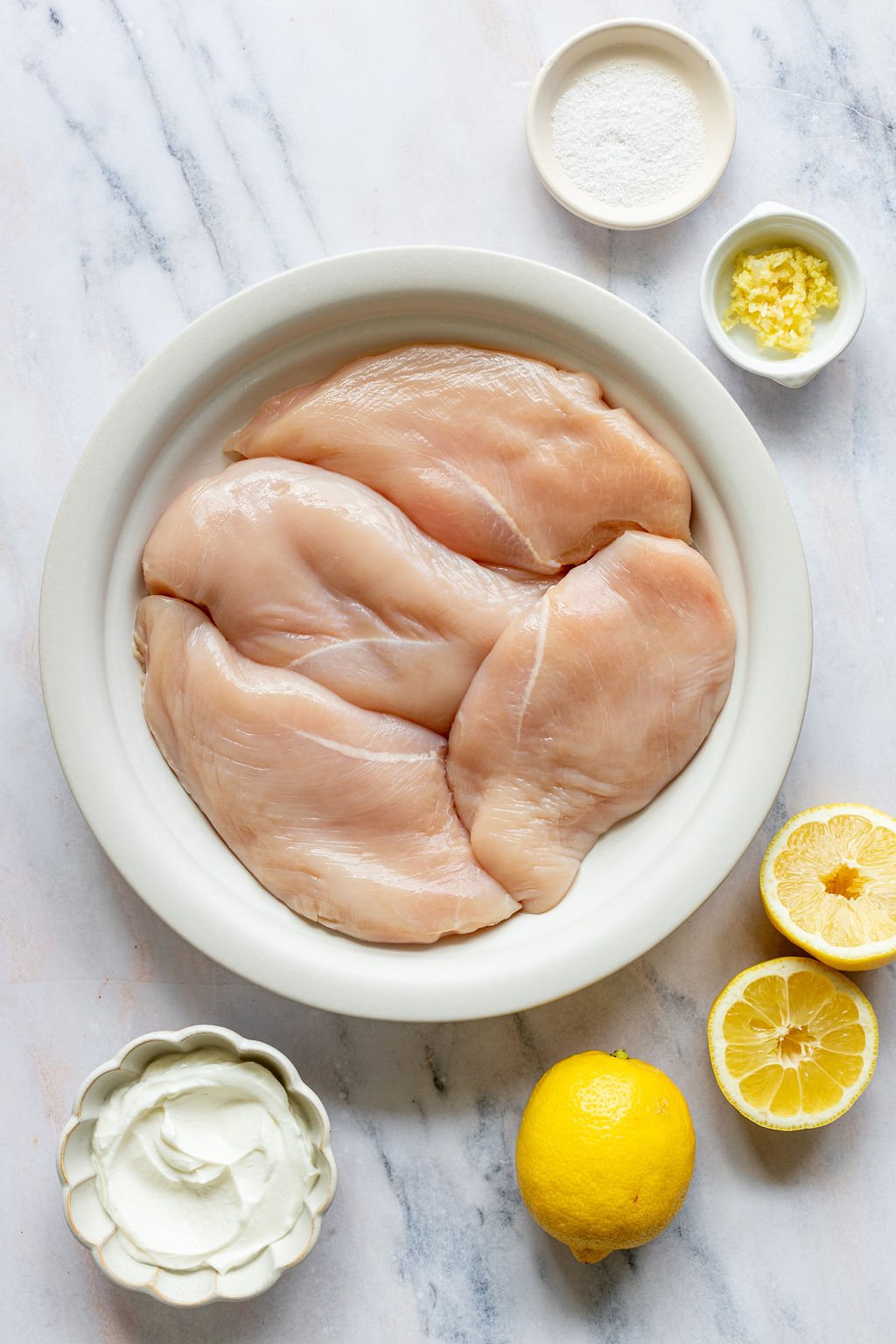 Overhead shot of the following ingredients: raw chicken breasts, yogurt, lemons, lemon zest, salt.