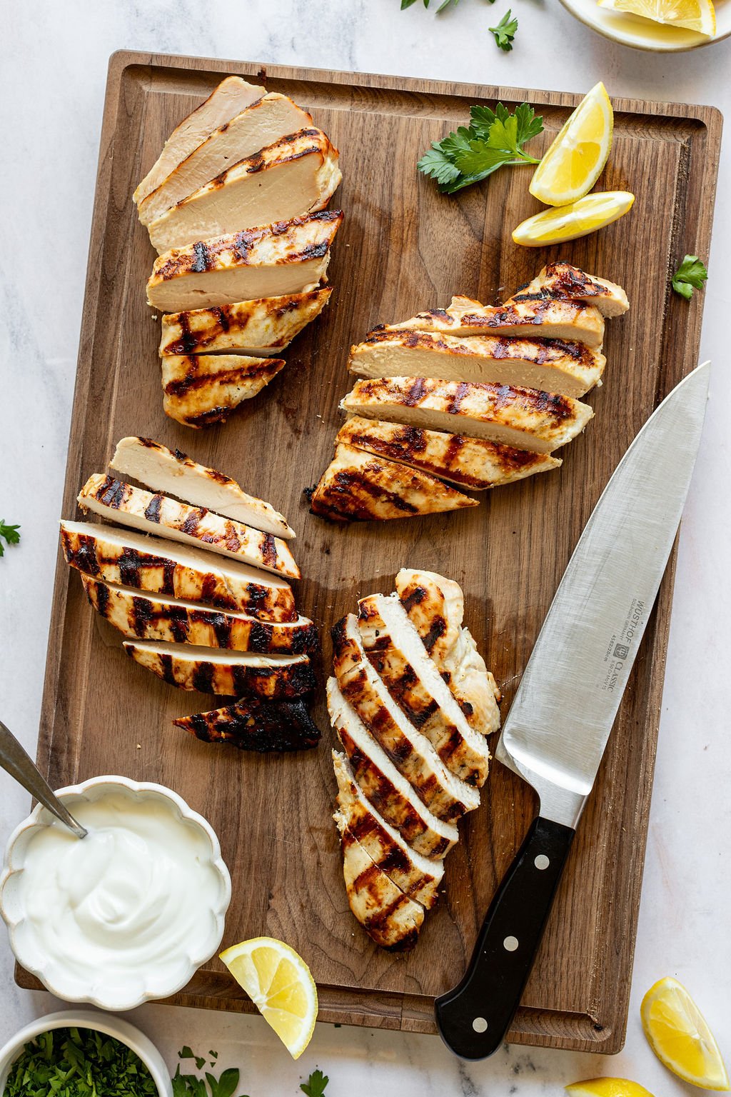 Overhead shot of a wooden cutting board with sliced grilled chicken breasts, lemon wedges, a knife, and a bowl of yogurt. Cutting board is surrounded by small bowls of chopped parsley and lemon wedges.
