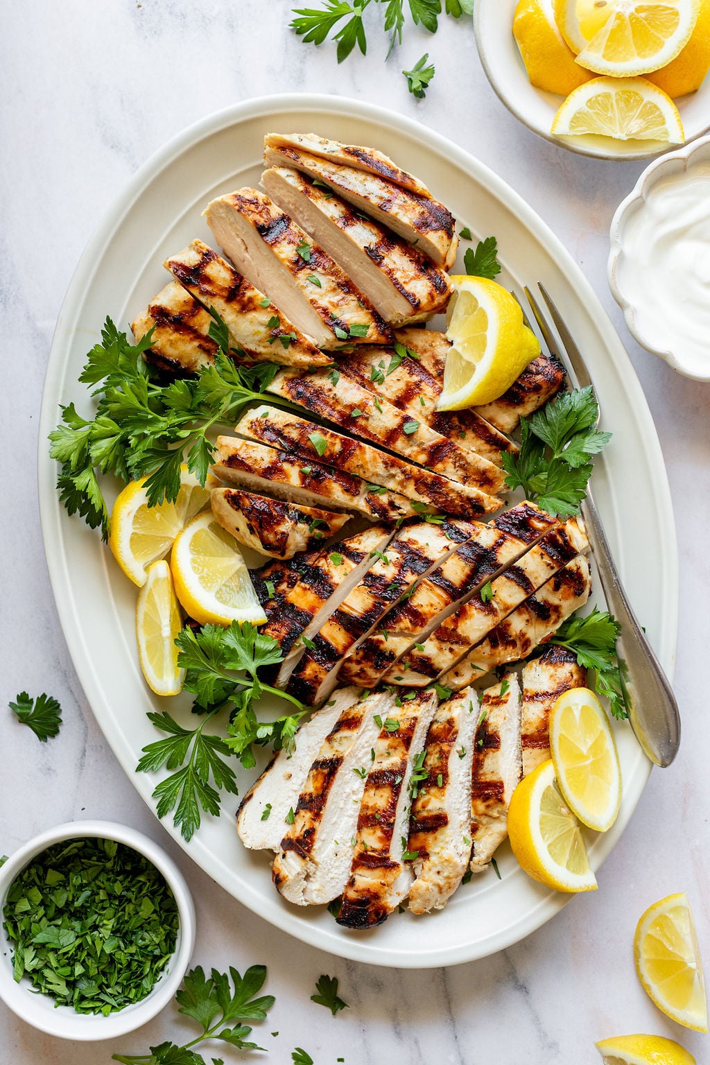 Close-up overhead shot of a white oval serving plate with sliced grilled chicken breasts, fresh parsley, and lemon wedges. Plate is surrounded by small bowls of chopped parsley, lemon wedges, and yogurt.