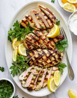 Close-up overhead shot of a white oval serving plate with sliced grilled chicken breasts, fresh parsley, and lemon wedges. Plate is surrounded by small bowls of chopped parsley, lemon wedges, and yogurt.