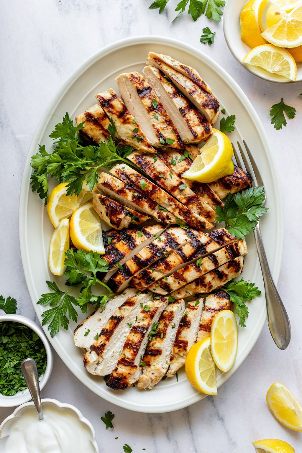 Close-up overhead shot of a white oval serving plate with sliced grilled chicken breasts, fresh parsley, and lemon wedges. Plate is surrounded by small bowls of chopped parsley, lemon wedges, and yogurt.