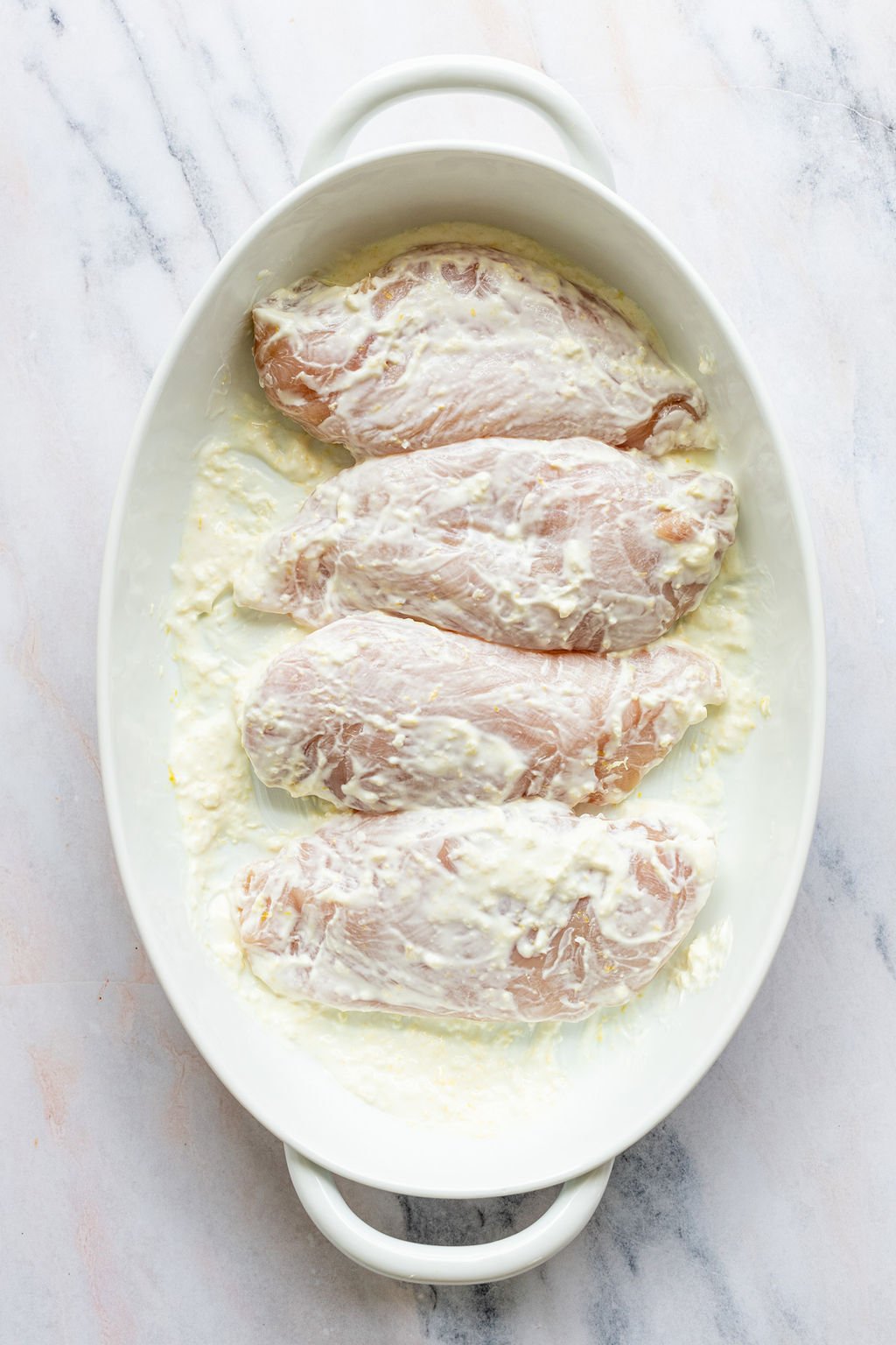 White oval baking dish with raw chicken breasts covered in white marinade.