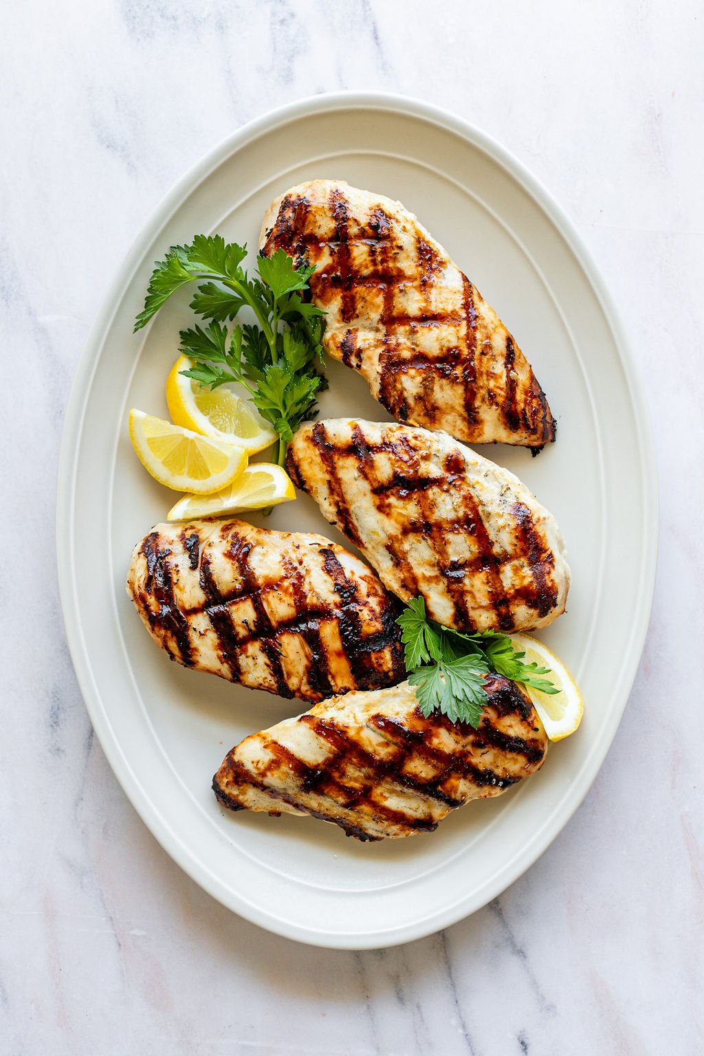 Overhead shot of a white oval serving plate with grilled chicken breasts, fresh parsley, and lemon wedges.