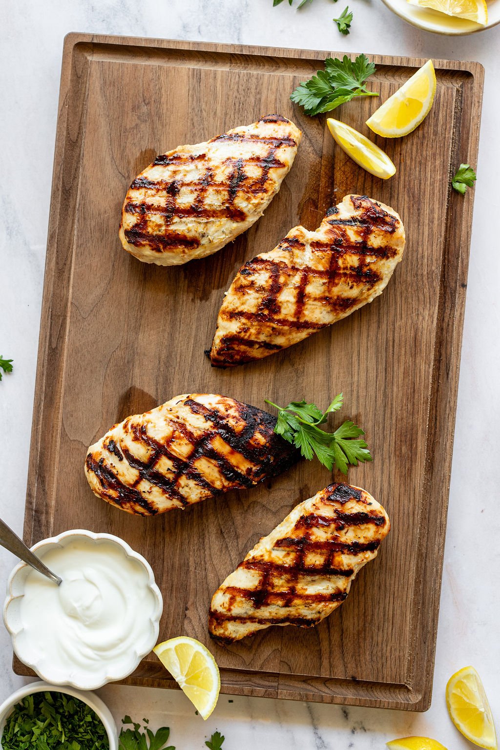 Overhead shot of a wooden cutting board with grilled chicken breasts, fresh parsley, lemon wedges, and a bowl of yogurt. Cutting board is surrounded by small bowls of chopped parsley and lemon wedges.