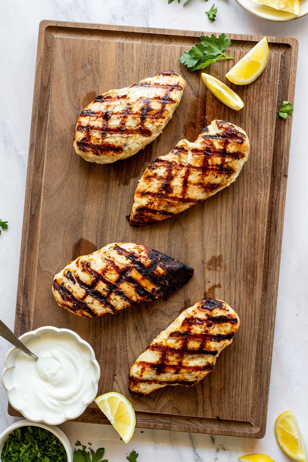 Overhead shot of a wooden cutting board with grilled chicken breasts, lemon wedges, and a bowl of yogurt. Cutting board is surrounded by small bowls of chopped parsley and lemon wedges.