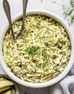 Close-up overhead shot of a white bowl with creamy coleslaw and servings spoons. Bowl is surrounded by a striped towel, pickle spears, and fresh dill.