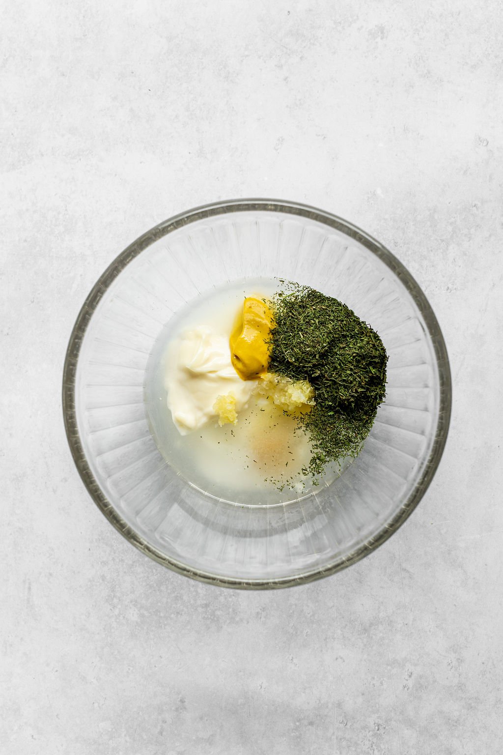 Overhead shot of a glass bowl with vinegar, mayonnaise, mustard, garlic, sugar, and dried dill.