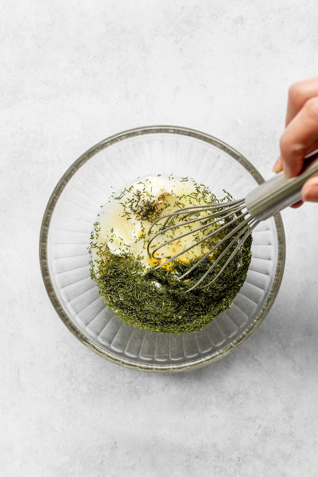 Overhead shot of whisking dried dill, vinegar, mayonnaise, and mustard in a glass bowl.