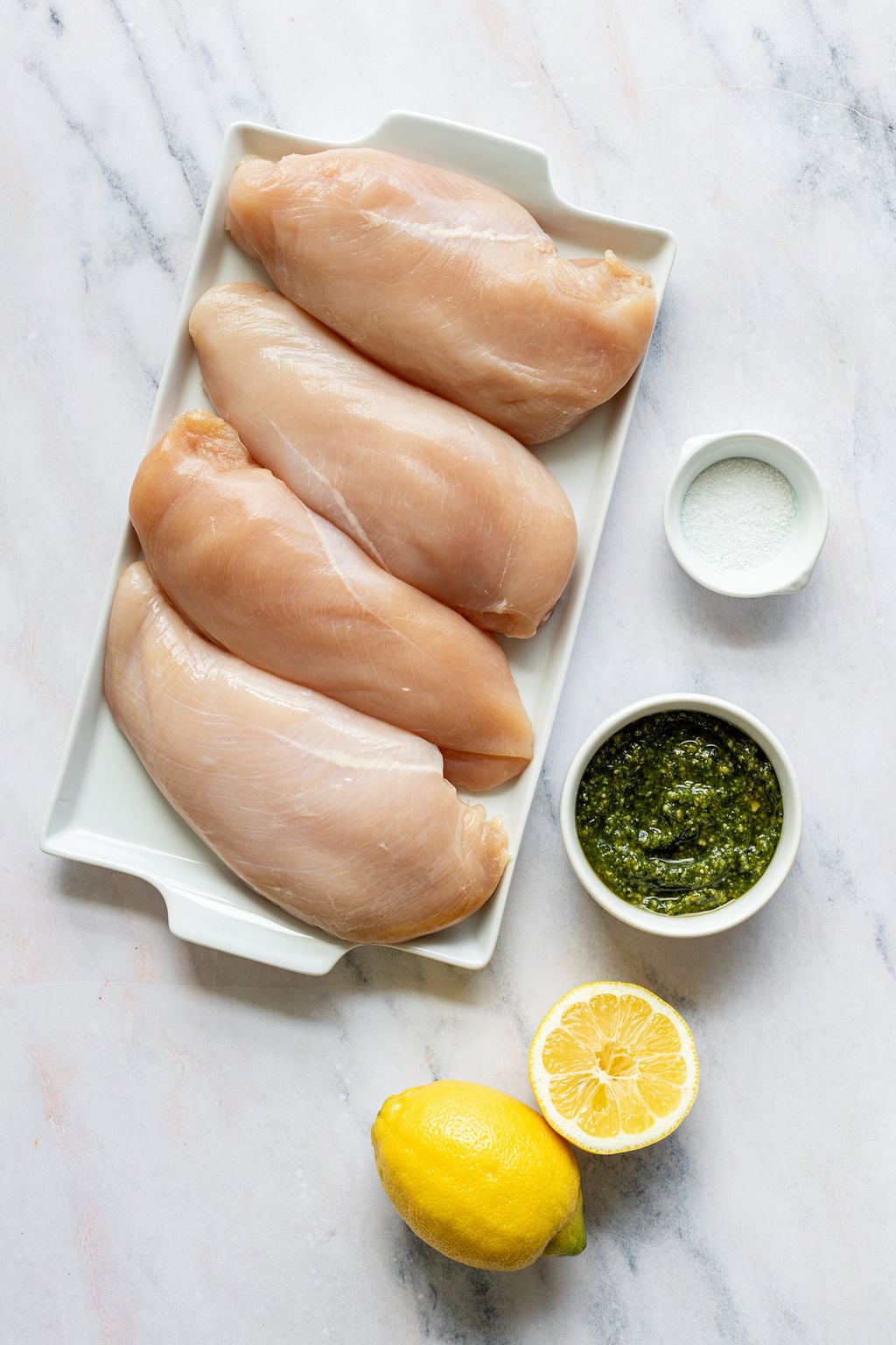 Overhead shot of the following ingredients: chicken breasts, salt, pesto, lemon.