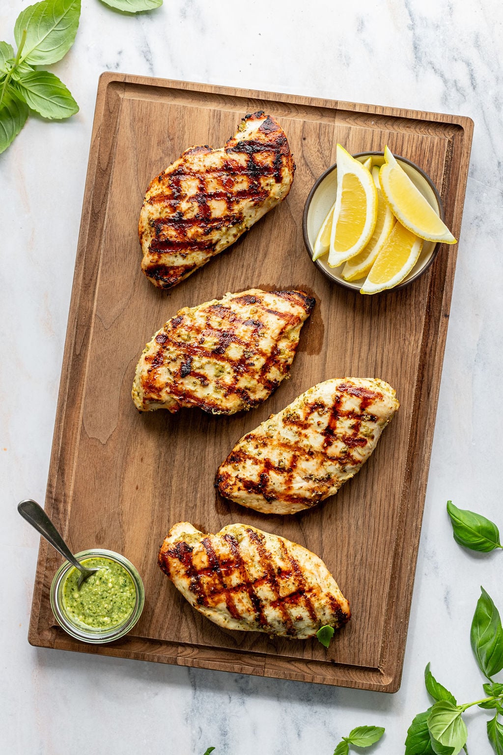 Overhead shot of a wooden cutting board with grilled chicken and small bowls of lemon wedges and green sauce. Cutting board is surrounded by fresh basil.