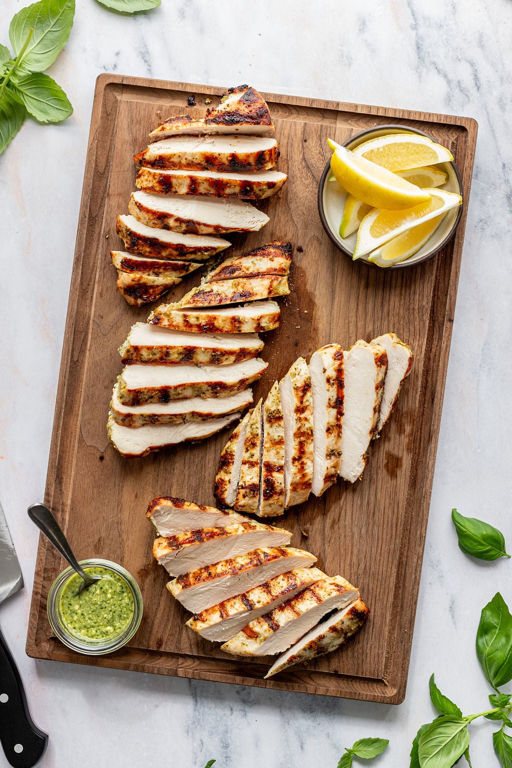 Overhead shot of a wooden cutting board with sliced grilled chicken and small bowls of lemon wedges and green sauce. Cutting board is surrounded by fresh basil.