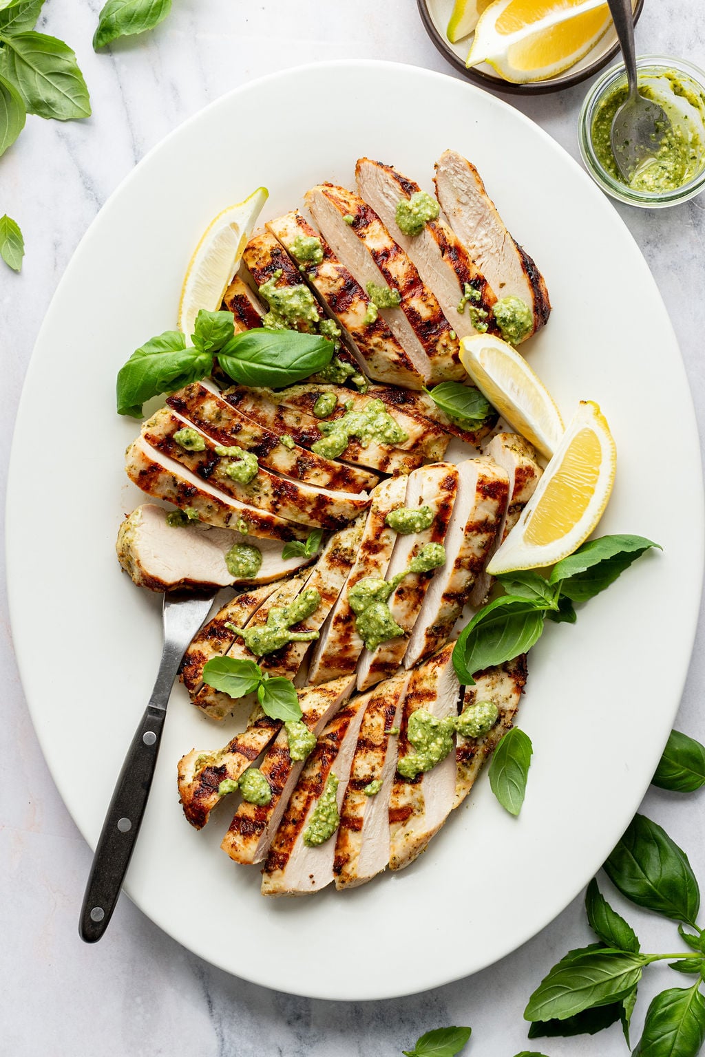 Close-up overhead shot of a white serving platter with sliced grilled chicken topped with green sauce, a serving fork, lemon wedges, and fresh basil. Platter is surrounded by fresh basil and a bowl of lemon wedges.
