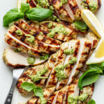 Close-up overhead shot of sliced grilled chicken topped with green sauce, fresh basil, lemon wedges, and a serving fork.
