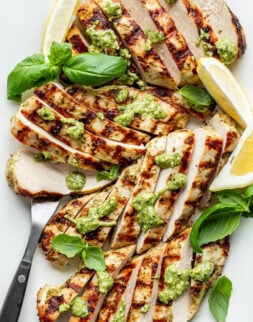 Close-up overhead shot of sliced grilled chicken topped with green sauce, fresh basil, lemon wedges, and a serving fork.