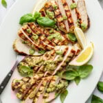 Close-up overhead shot of a white serving platter with sliced grilled chicken topped with green sauce, a serving fork, lemon wedges, and fresh basil.
