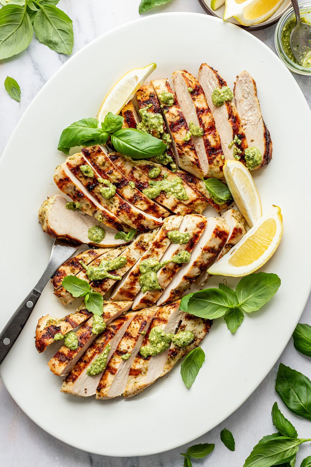 Close-up overhead shot of a white serving platter with sliced grilled chicken topped with green sauce, a serving fork, lemon wedges, and fresh basil.