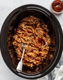 Close-up overhead shot of a crockpot with a fork and shredded meat covered in red sauce. A small bowl of red sauce and a spoon are in the corner.