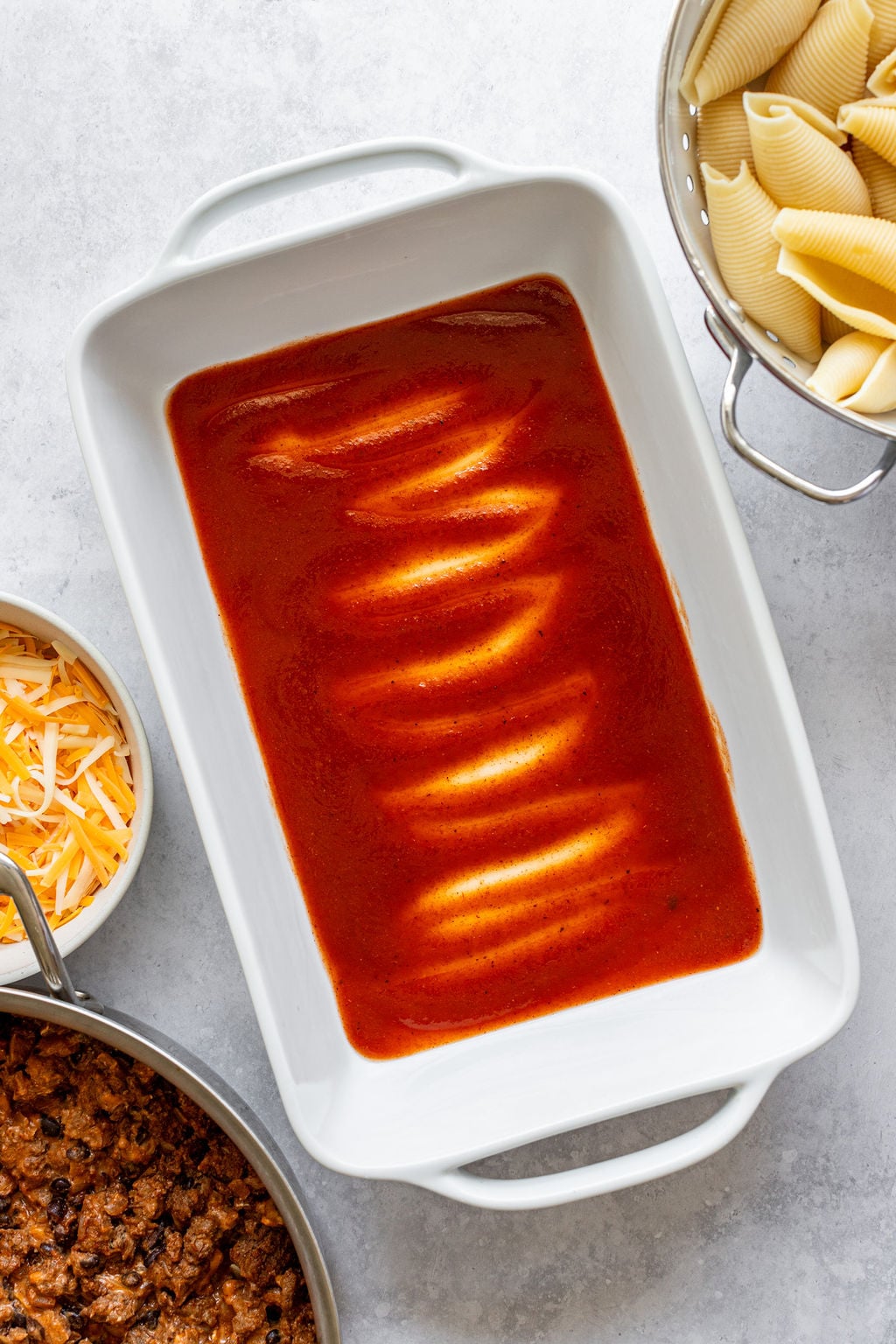 Baking dish with red sauce. Baking dish is surrounded by a bowl of shredded cheese, colander of cooked pasta shells, and saucepan of cooked ground beef.
